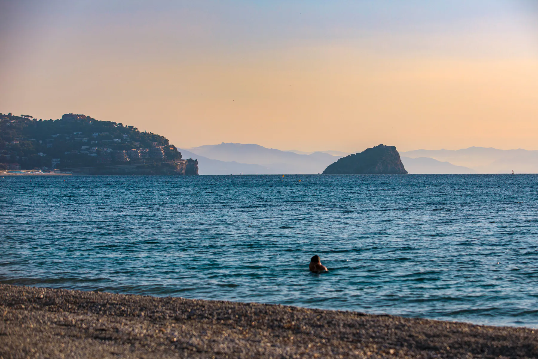 Una persona fa il bagno nel mare calmo al tramonto, all’orizzonte l’Isola di Bergeggi