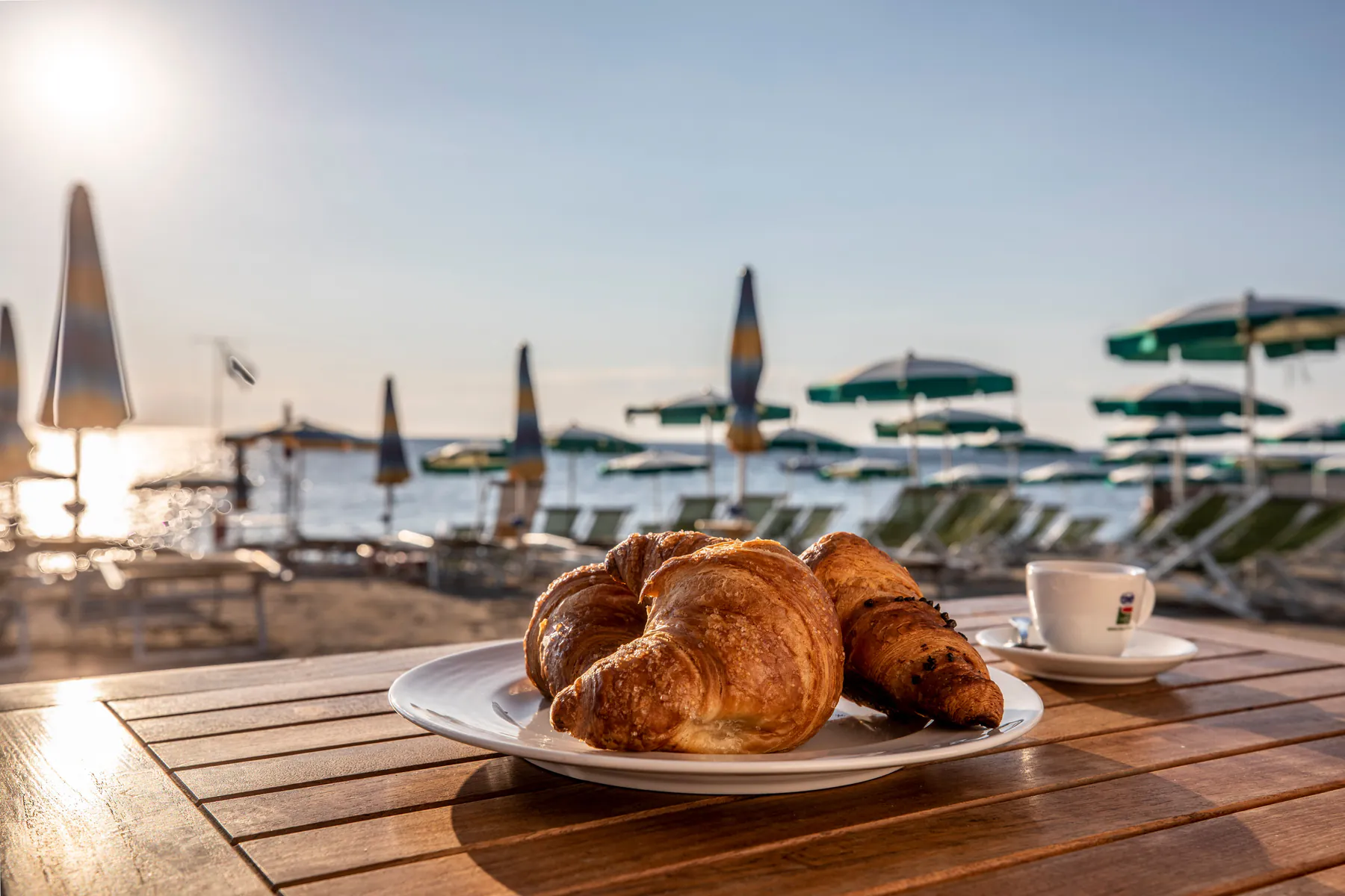 Croissant e caffè su un tavolino in spiaggia, sullo sfondo ombrelloni e mare